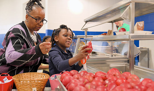 child in lunch line holding a pear